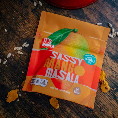 It's Nola Sassy Mango Masala package is placed against a wooden background with various seeds and dried mango slices. 