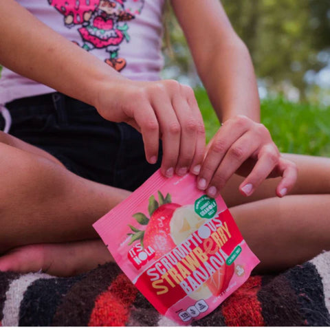 It's Nola Scrumptious Strawberry Banana is being opened by a young girl. The young girl is at the park and is seated on a black, red, and white striped blanket. 