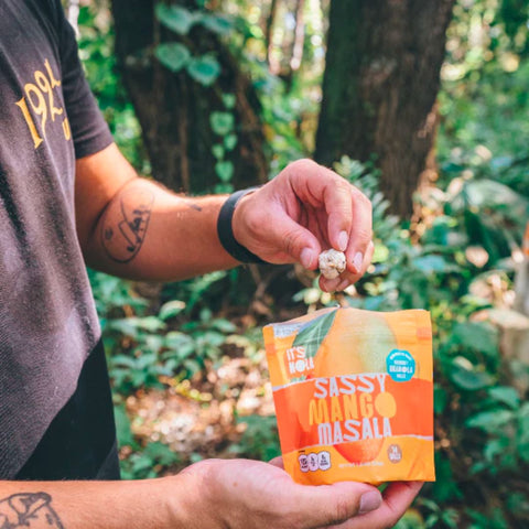 It's Nola Sassy Mango Masala package being held by a person who is about to reach for a yummy granola bite. They are eating their It's Nola in a forest with nature surrounding them.