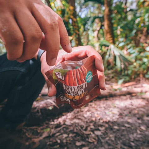 It's Nola Decadent Chewy Chocolate package being held by a person who is about to reach for a yummy granola bite. They are eating their It's Nola in a forest with nature surrounding them. 