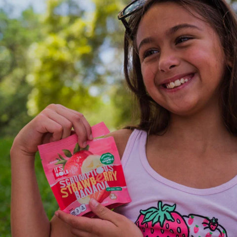 It's Nola Scrumptious Strawberry Banana is being held by a smiling young girl. She is happy to eat her Scrumptious Strawberry Banana.