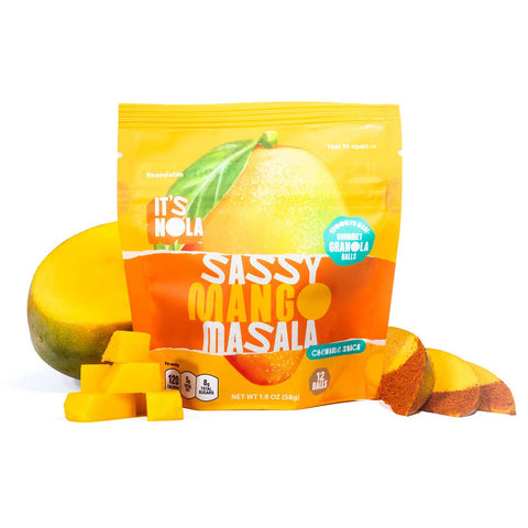 It's Nola Sassy Mango Masala package is surrounded by a fresh mango cut in half behind the package, mango cubes to the left of the package, and mango slices with spices to the right of the package. 