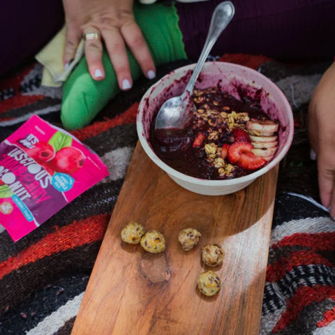 It's Nola Luscious Cranberry Coconut package is laying on a patterned blanket to the left of a person who is enjoying an acai smoothie bowl with It's Nola granola snack bites and sliced strawberries and bananas.