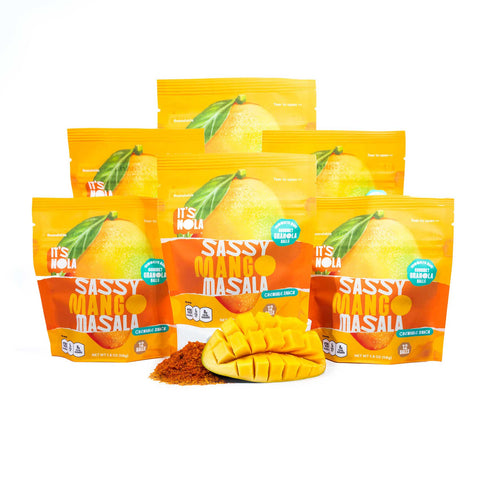 It's Nola Sassy Mango Masala is being placed in a pyramid with a sliced mango and spices under the center of the pyramid. The products are placed against a white background.  